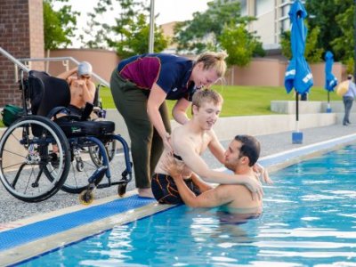 A young man is being helped from a wheelchair into a swimming pool by two trainers.